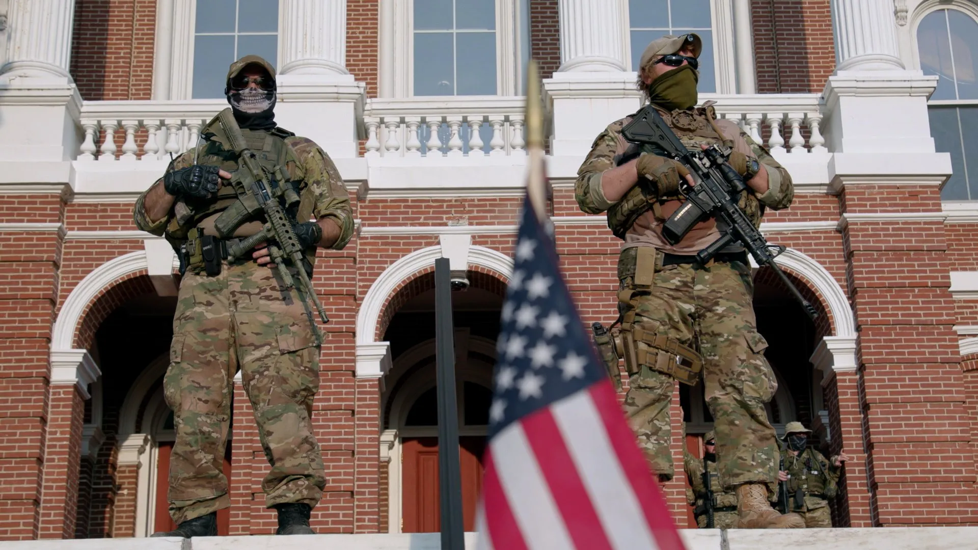 Two armed militia members stand guard outside a courthouse with an American flag